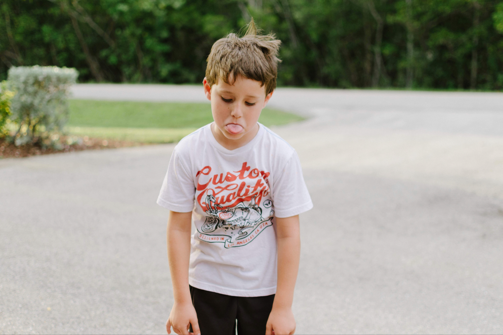 Bored boy in the garden, with tongue out, black pants and white t-shirt with red and black letters.