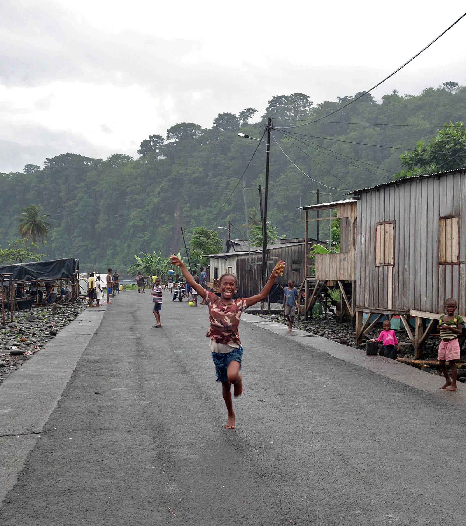 Criança africana a correr numa estrada, com casas de madeira laterais.