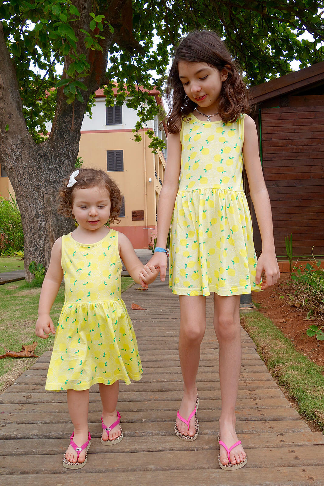 Meninas de vestido amarelo a caminharem de mãos dadas sobre um passadiço de madeira.