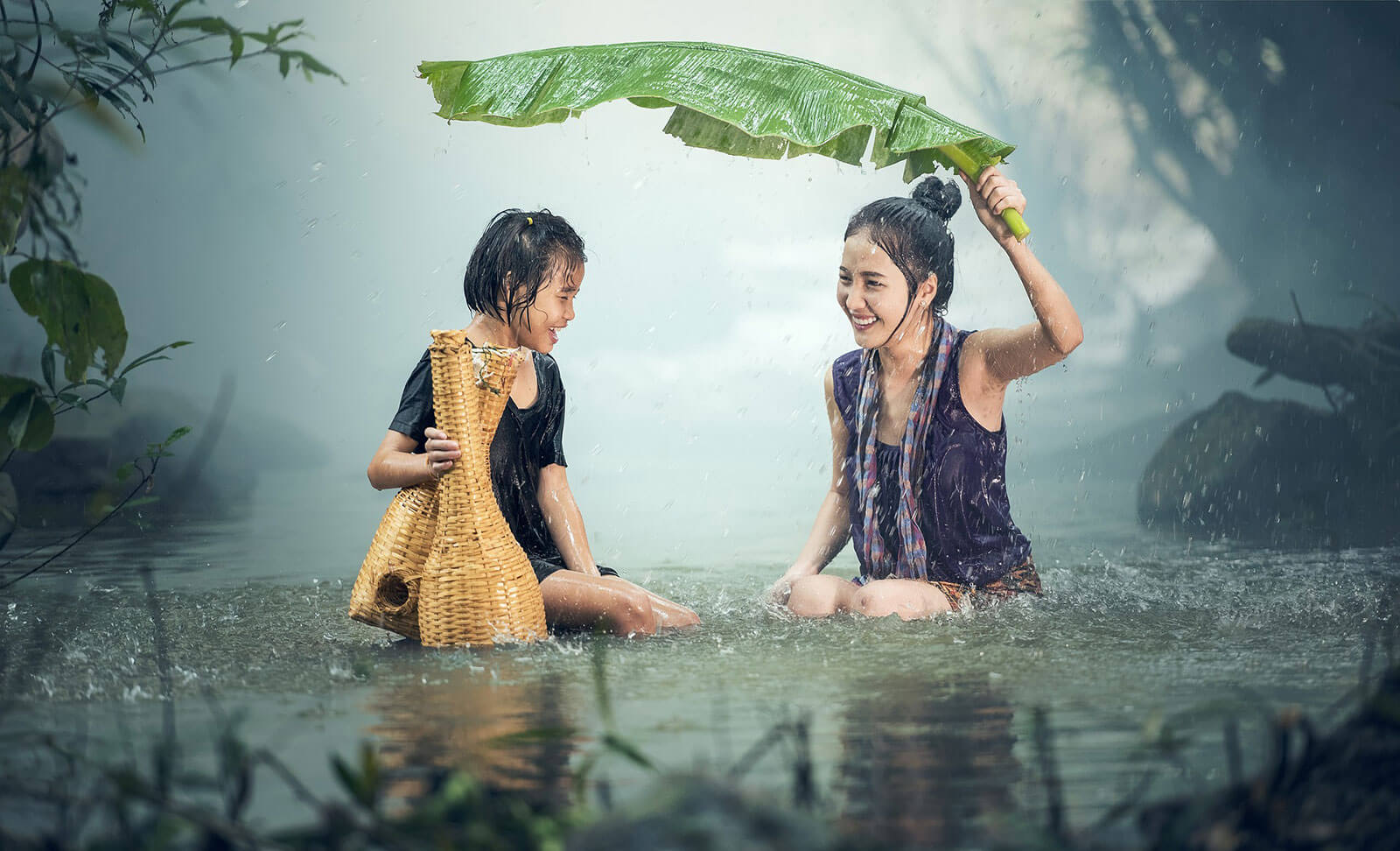 Mulheres asiáticas dentro de um rio, protegidas da chuva com folha de bananeira.