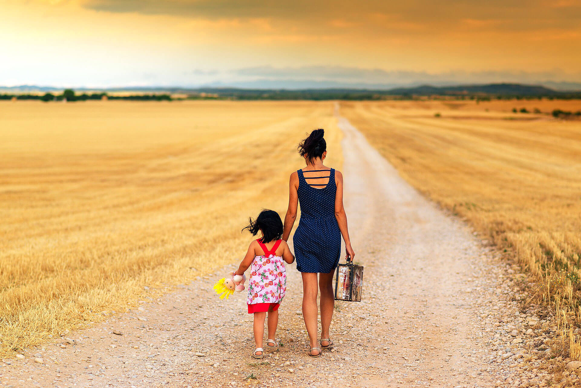 Mãe que transporta mala e criança com boneco na não, que caminham de mãos dadas através de um caminho de terra numa seara.