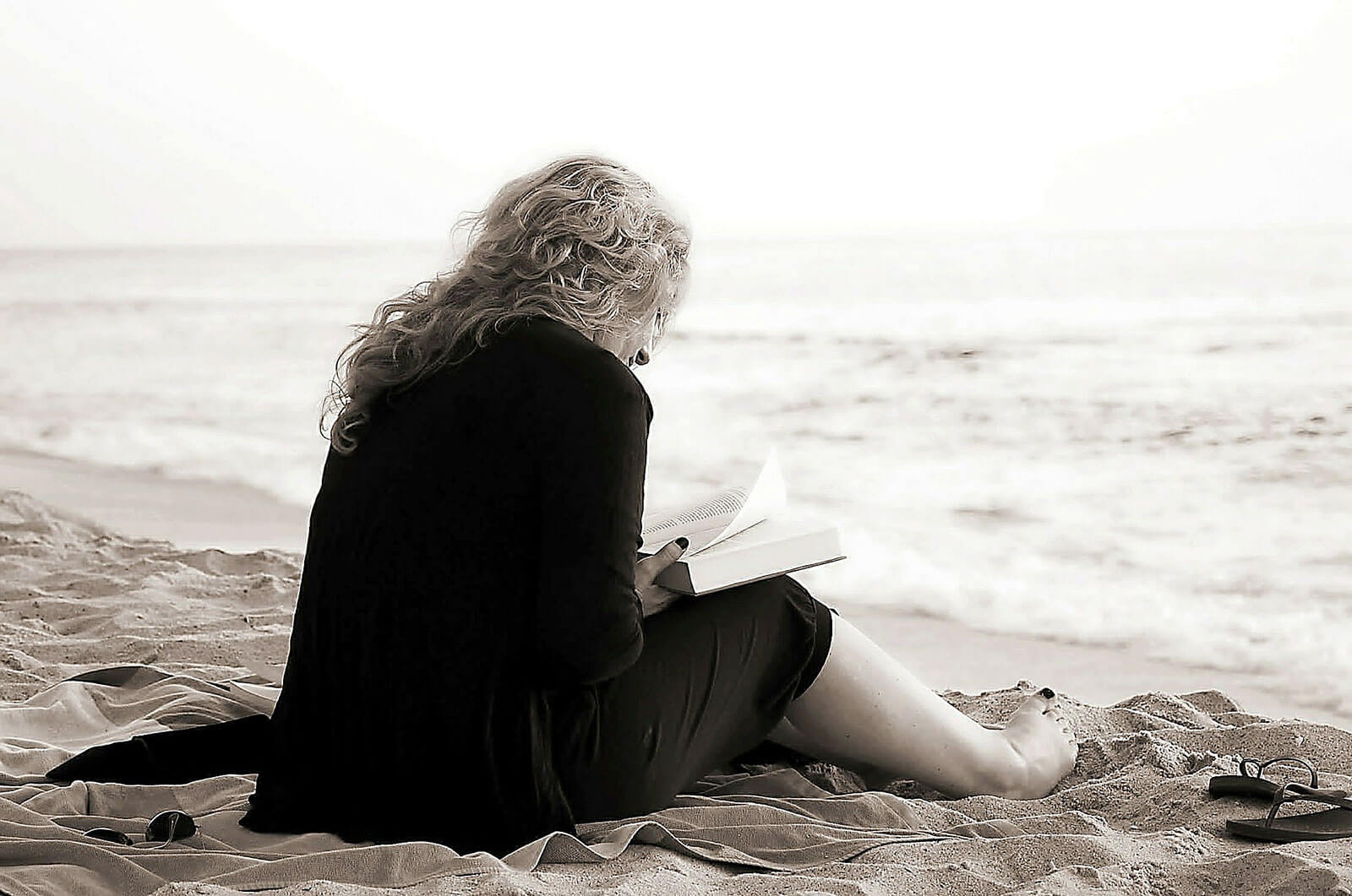 Mulher a ler um livro, sentada na areia, junto ao mar.