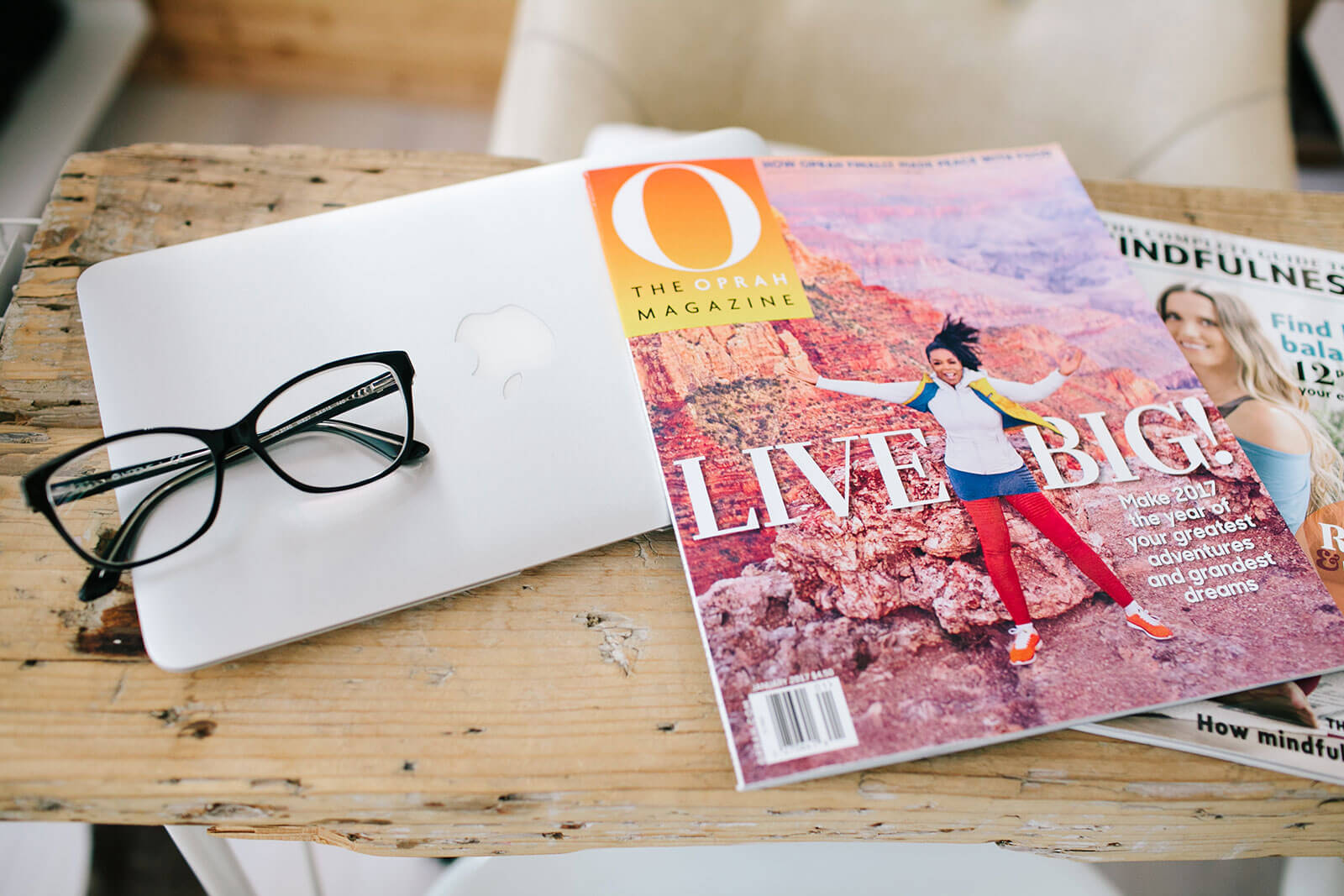 Revista Oprah, computador portátil e óculos sobre uma mesa de madeira.