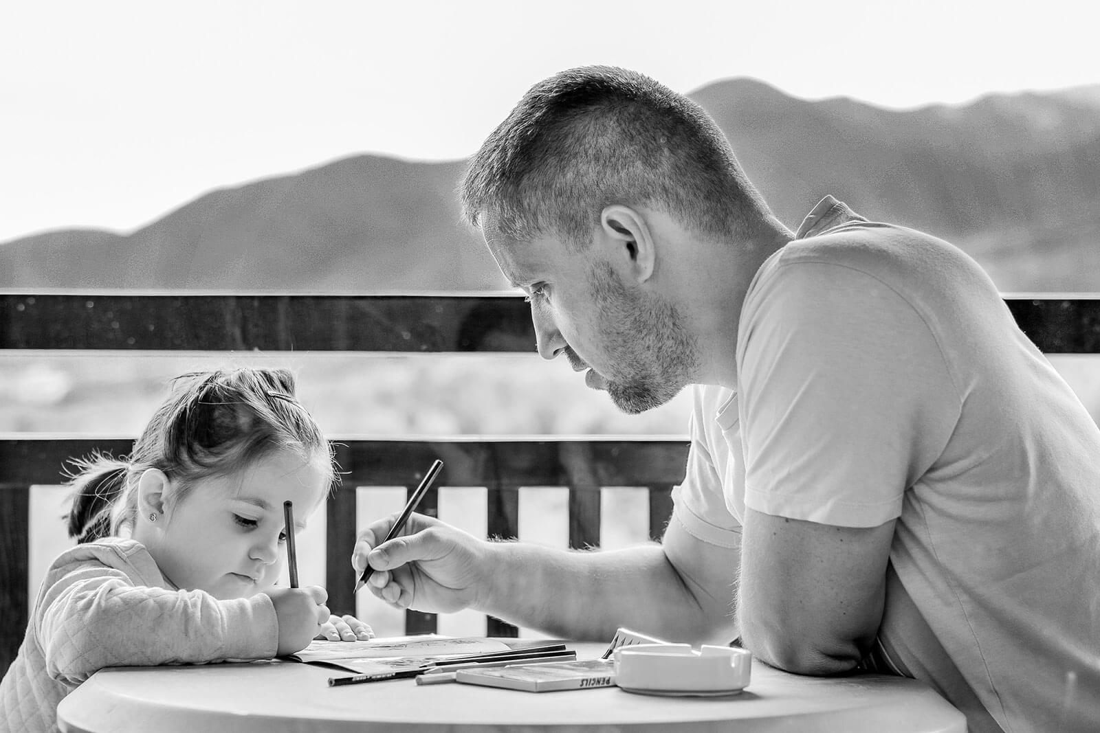 Pai e filha sentados à mesa, numa varanda, a escrever em cadernos.