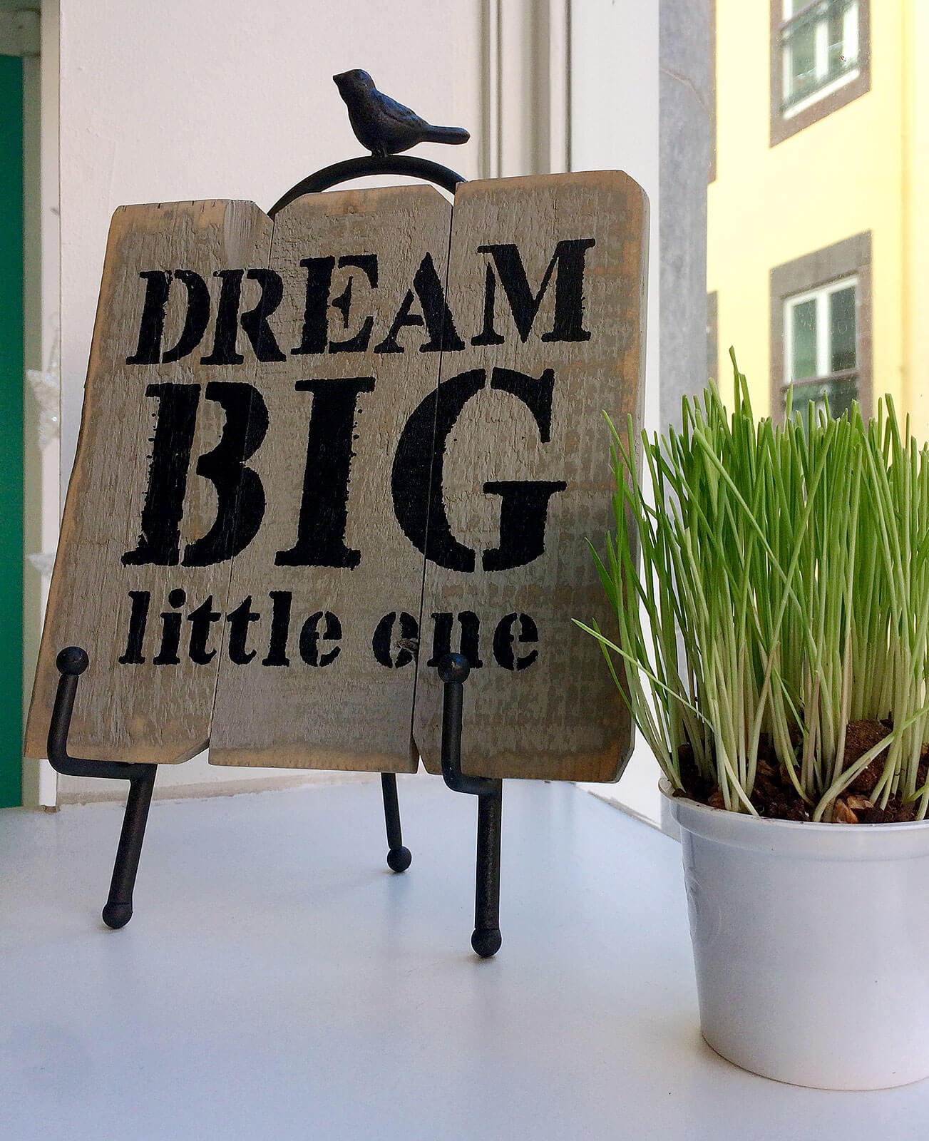 Placard com texto "Dream Big little one" e vaso com planta junto a uma janela.