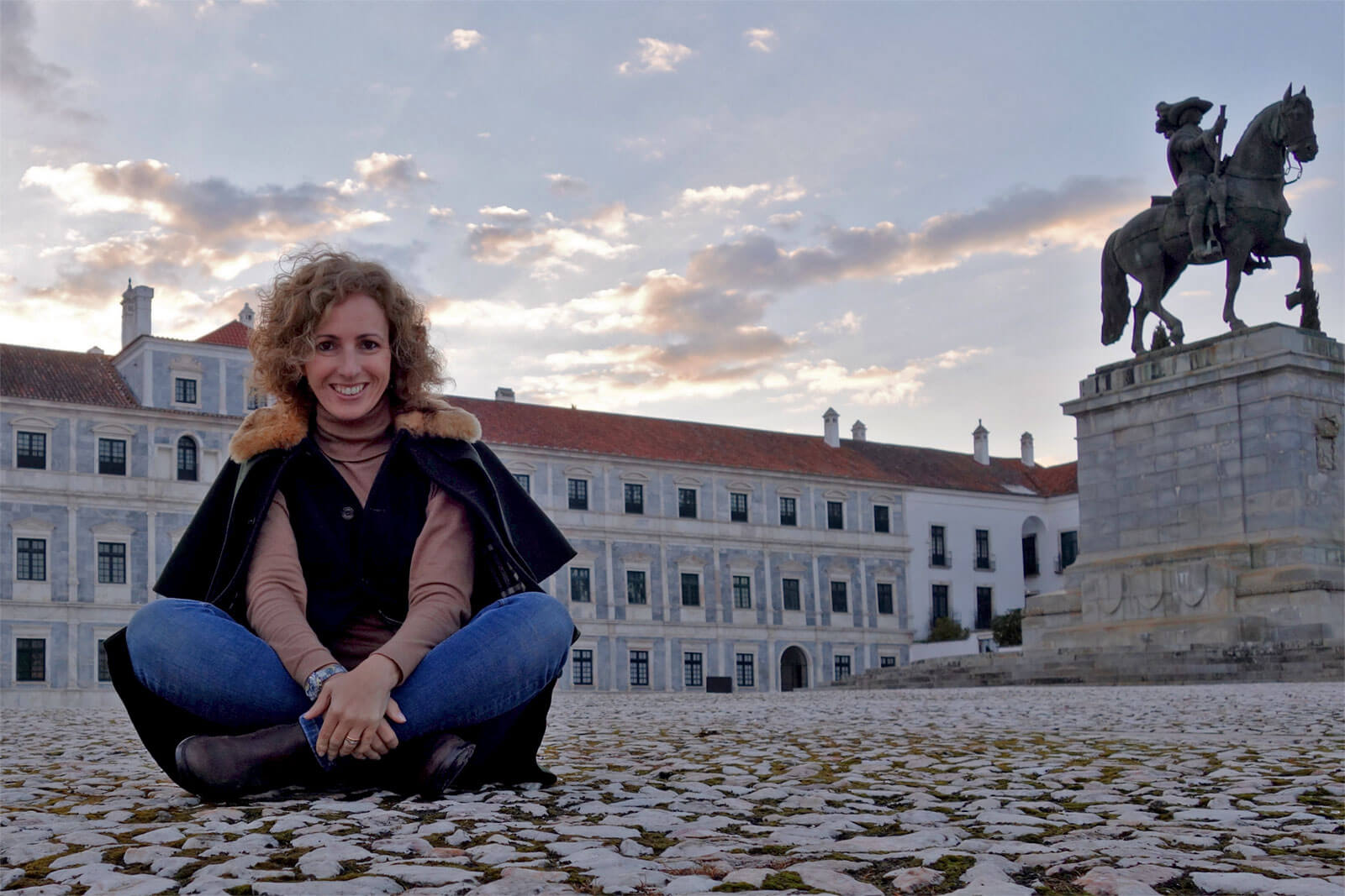 Rita Aleluia sentada numa grande praça, com pavimento em pedra rústica e grande estátua de cavaleiro.