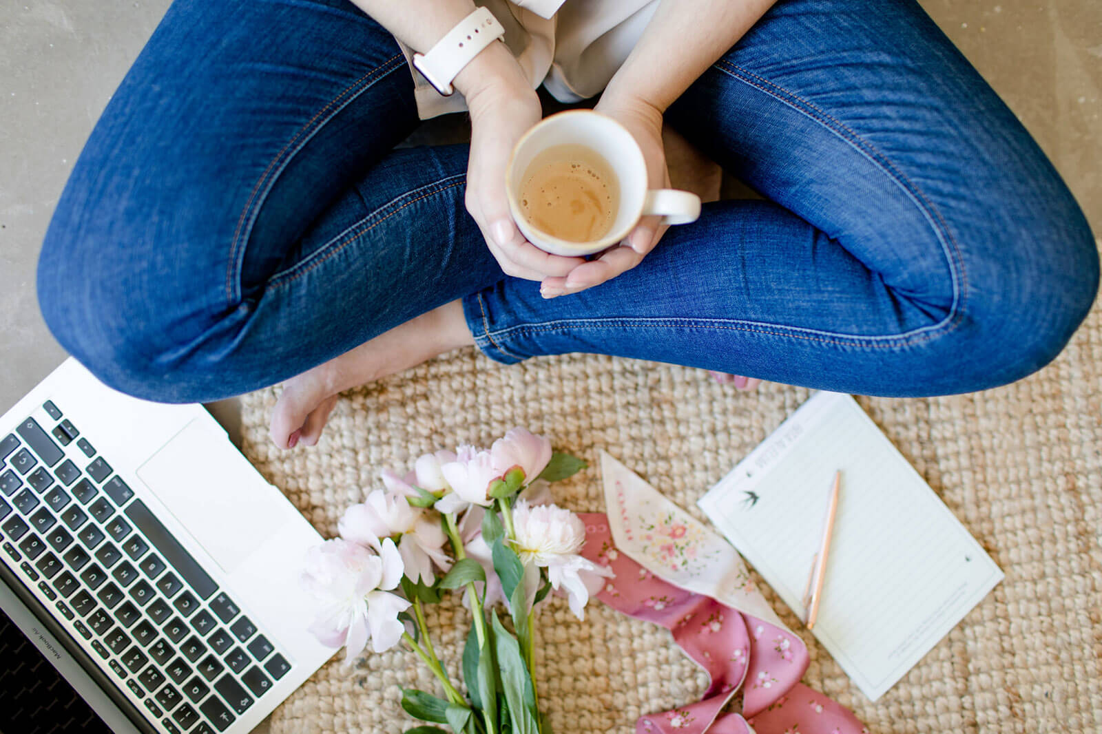Mulher de pernas cruzadas a segurar numa chávena de café, junto a computador portátil e flores.