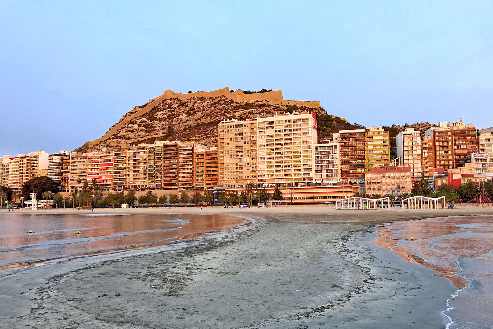 Linha de prédios urbanos entre praia e monte circundado por muralha, na cidade de Alicante.