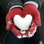 Heart made of white snow, resting in hands covered with red gloves, sprinkled with snow, in a half body covered with a black coat and a gray sweater.